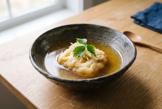 Kumo-no-Ochishizuku: Cloud-Drop Tofu in Golden Dashi A single golden-fried fluffy tofu cloud served in a dark ceramic bowl with clear dashi broth and green herbs on a minimalist wooden table.