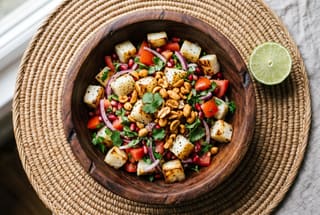 Charred Cassava and Berbere Groundnut Kachumbari A vibrant salad of charred cassava, tomatoes, onions, and spiced peanuts in a wooden bowl on a woven mat with lime slices.