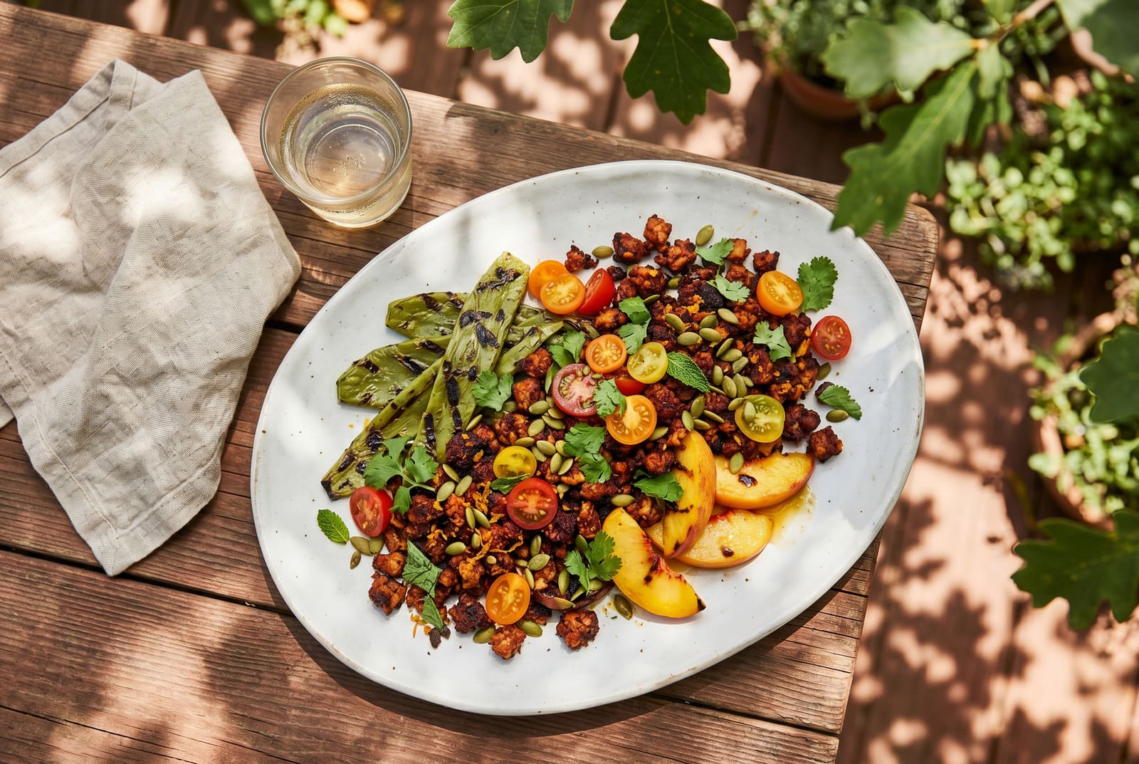 Vibrant guajillo tempeh crumble with charred nopales and peaches on a white platter, set on a sunlit redwood table in a California garden.