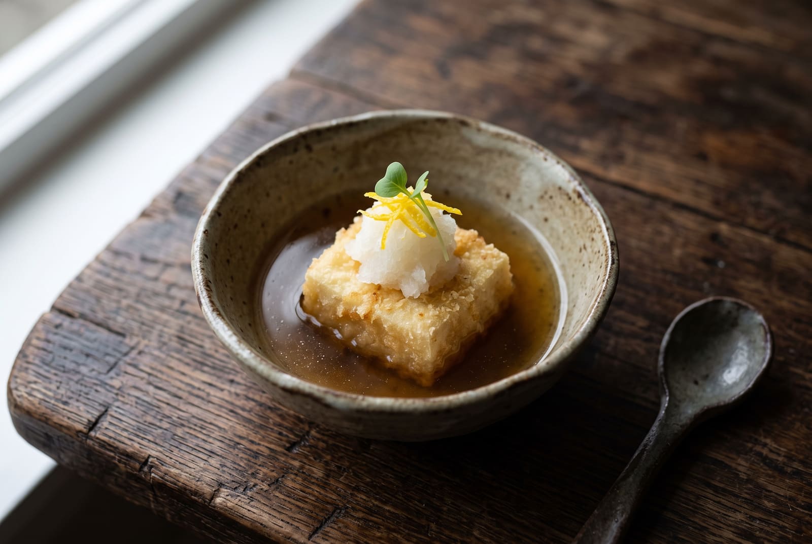 Binchotan-Seared Agedashi Tofu in Yuzu-Kombu Shizuku Crispy agedashi tofu in clear golden dashi, topped with grated daikon and yuzu zest in a rustic ceramic bowl.