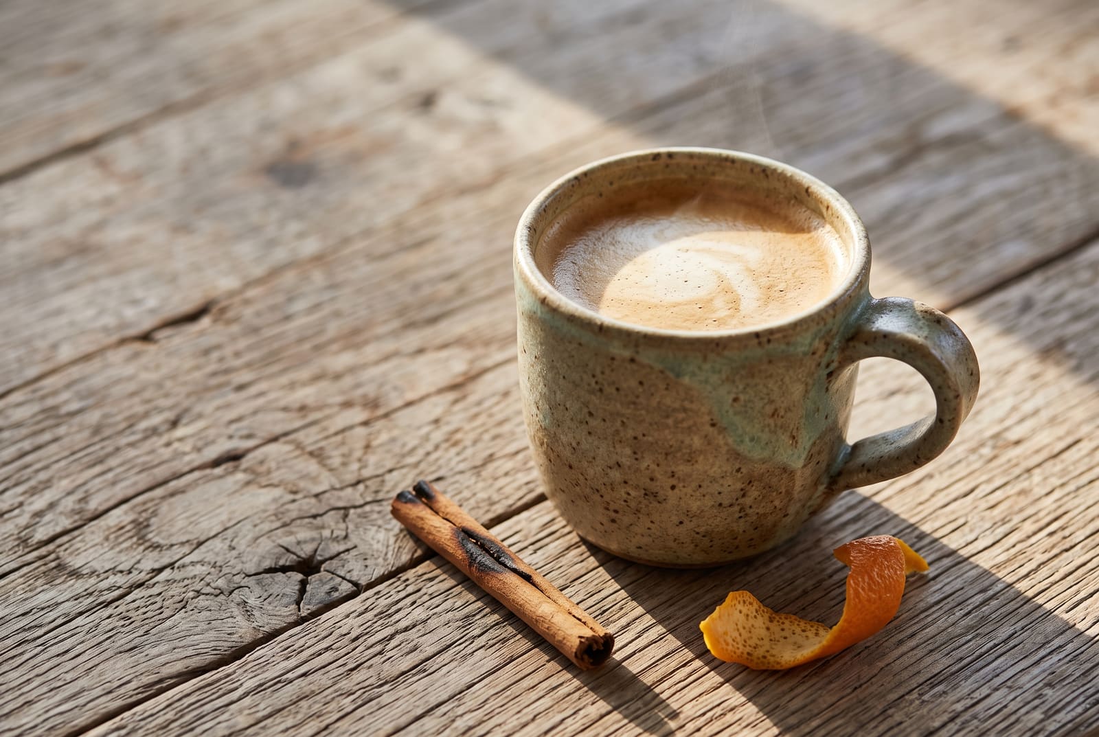 A rustic ceramic mug of frothy steamed coffee garnished with orange zest and dark chocolate shavings on a sunlit wooden table.