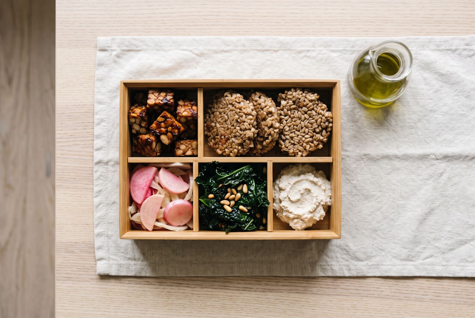 A wooden bento box filled with balsamic glazed tempeh, fermented farro cakes, colorful pickled vegetables, wilted kale, and cultured ricotta.