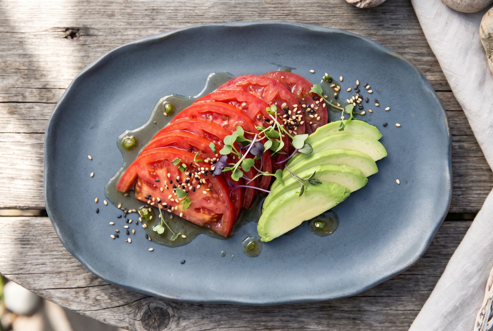 Thinly sliced ruby red confit tomato sashimi garnished with avocado, sesame seeds, and ponzu on a slate ceramic plate.