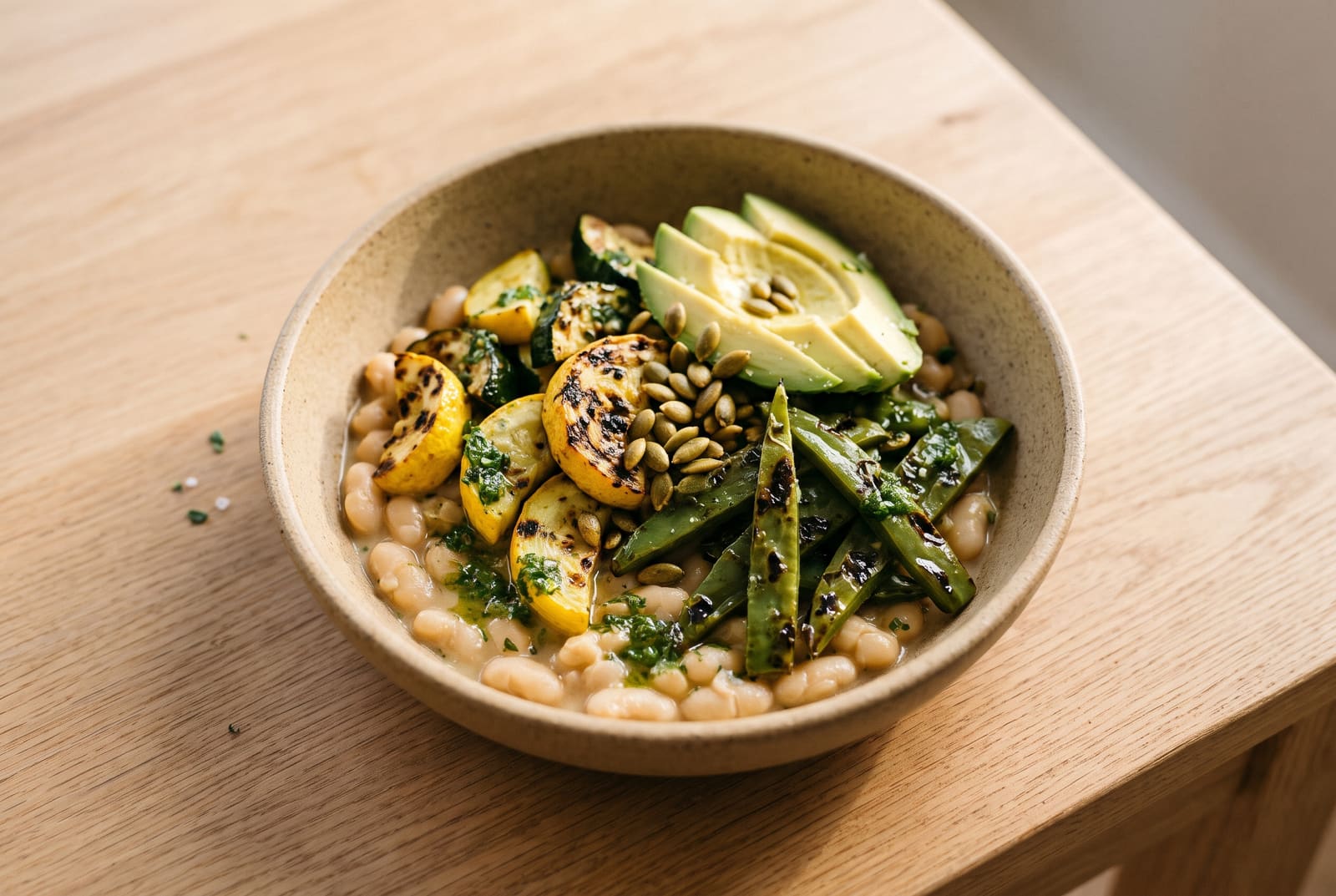 Charred squash and nopales served over creamy white beans, topped with green herb mojo and avocado in a rustic ceramic bowl.