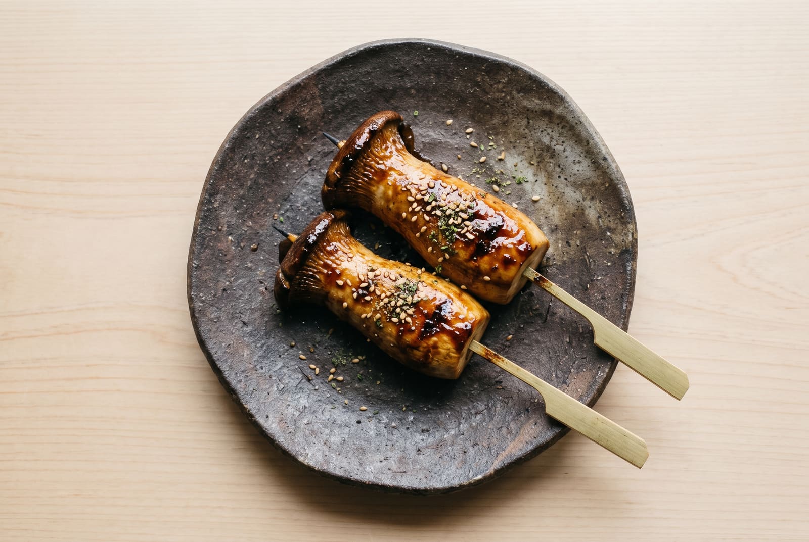 Binchotan-grilled king oyster mushroom skewers with glistening caramelized miso glaze on a dark wabi-sabi stoneware plate.