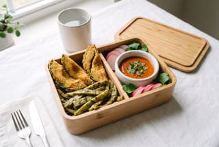 A wooden bento box with crispy fried avocado and cactus strips, a small bowl of red sauce, and radish garnishes on a white cloth in bright sunlight.