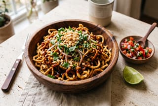 Mombasa Sunset Crispy Noodles with Tamarind-Coconut Glaze Crispy golden noodles with dark tamarind glaze, crushed peanuts, and fresh cilantro in a wooden bowl on a clean table.