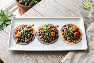 Grilled Nopales and Al Dente Heirloom Bean Tostaditas Three vegan tostaditas topped with heirloom beans, grilled nopales, and tomatoes on a white plate under soft, natural sunlight.
