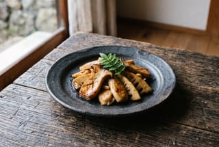 Binchotan-Scented King Trumpet Mushrooms with Young Bamboo Sautéed glazed king trumpet mushrooms and bamboo shoots on a dark ceramic plate with a single green leaf garnish.