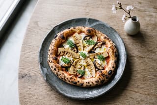 Shojin Sourdough Pizza with Binchotan-Charred Takenoko and White Miso Cream Artisan vegan pizza with charred bamboo shoots and green leaves on a dark rustic ceramic plate.