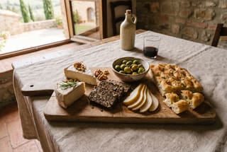 A rustic wooden board with vegan cashew cheese, lentil terrine, olives, sliced pears, and focaccia on a linen tablecloth.