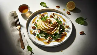 Image of a syrupy Asian pear salad on a white plate, with pears in a spiral, walnuts, vegan feta, mint, sesame seeds, and a glossy dressing, on a simple table setting.