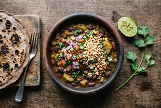 A clay bowl of dark bean and green banana stew topped with fresh tomato-onion salad and crushed peanuts on a wooden table.