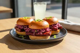 Three sticky Gochujang seitan sliders with kimchi slaw on a matte black plate, set on a sunlit oak table. Vibrant Asian-American vegan BBQ fusion.