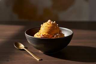 Vibrant ochre pumpkin ice cream topped with popped sorghum in a dark ceramic bowl, illuminated by warm golden sunlight.