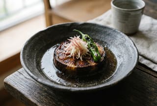 Binchotan-Seared Kamo Nasu in Chilled Kombu-Tamari Dashi Seared Kyoto eggplant round topped with ginger and shishito pepper in a handmade ceramic bowl on a dark wood table.
