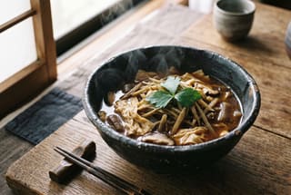 Kaze no Kaori Shredded Root and Yuba Stew A steaming bowl of shredded root and yuba stew in a dark ceramic bowl on a wooden table, garnished with shiso.