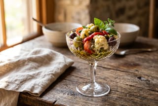 Vibrant marinated cauliflower, olives, and red peppers in a crystal glass on a rustic wooden table under soft natural light.