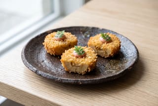 Three golden breaded bamboo medallions on a dark ceramic plate, topped with pink sauce and a green leaf, in a minimalist Japanese setting.
