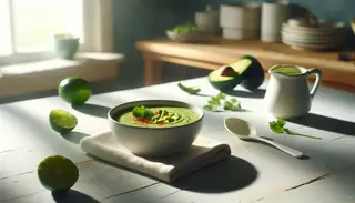 Creamy vegan cold soup in white bowl on wooden table, highlighted by natural light, with cilantro and chili flakes garnish, beside spoon and lime slice.