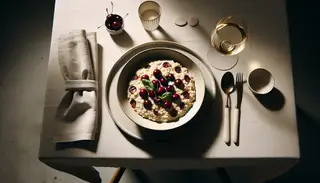A minimalist dinner table featuring a creamy vegan sour cherry risotto in a shallow bowl, accented with fresh basil and served with white wine under warm lighting.