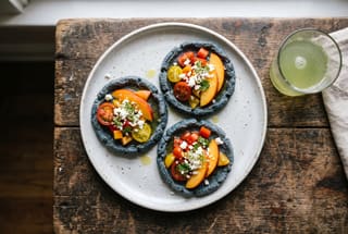 Smoked Blue Masa Sope with Heirloom Tomato and Nectarine Aguachile Three blue corn sopes topped with colorful tomato and nectarine aguachile on a ceramic plate, set on a rustic wooden table in soft sunlight.