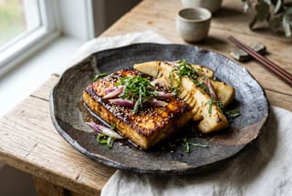 Binchotan-Seared Silken Tofu & Spring Bamboo Street Steak Pan-fried silken tofu and bamboo shoots with dark miso glaze on a dark ceramic plate, topped with fresh shiso and myoga ginger.