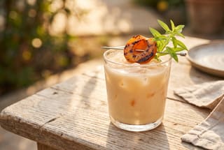 Charred Apricot & Toasted Almond Horchata A glass of creamy apricot horchata garnished with a charred fruit slice and green herbs on a sunlit wooden table.