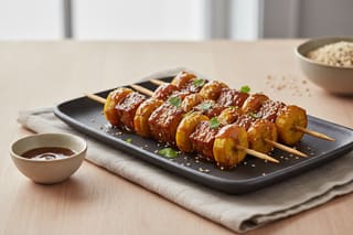 Glossy miso-tamarind glazed plantain and tempeh skewers with sesame seeds and cilantro on a dark platter. High-end African-Asian vegan fusion.
