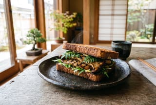 The Binchotan-Seared Eringi & Smoked Miso Flambé A binchotan-grilled king oyster mushroom sandwich on a dark ceramic plate, garnished with green leaves and served on a minimalist wooden table.