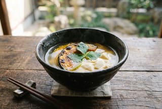 A minimalist dark ceramic bowl of Japanese kabocha and tofu soup on a wooden table in soft natural light.