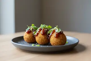 Crispy Suya plantain croquettes with dark miso-tamarind glaze, sesame, and scallions on a charcoal plate. Minimalist high-end vegan tapas.