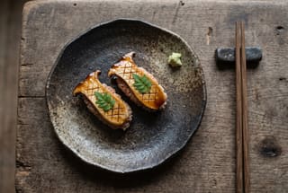 Binchotan Charred King Oyster Zuke Nigiri with Aged Akazu Rice Two pieces of charred king oyster mushroom zuke nigiri on a dark handmade ceramic plate with a tiny dab of fresh wasabi.