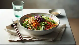 Vibrant vegan Asian-inspired salad in a white ceramic bowl, featuring quinoa, colorful veggies, and sesame seeds, with chopsticks and water glass on a simple table.