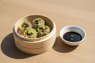 Sunlit vegan Porcini and Edamame Siu Mai in a bamboo steamer, garnished with Thai basil and served with balsamic-soy glaze on a light oak table.