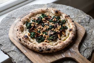 Tuscan Cavolo Nero and Wild Mushroom Artisan Pizza Artisan sourdough pizza topped with white cannellini cream, wilted cavolo nero, seared wild mushrooms, and walnuts on a wooden peel.