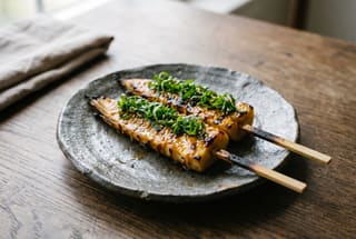 Binchotan-Grilled Takenoko with Young Shiso Pesto Charred bamboo shoot skewers with a glossy miso glaze and green shiso pesto on an artisanal grey plate.
