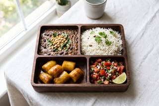 A vegan Swahili bento box with black bean stew, coconut rice, tamarind green bananas, and fresh kachumbari on a white tablecloth.