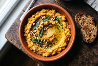 A textured golden squash and bean mash topped with sage and hazelnuts in a rustic bowl on a wooden table.