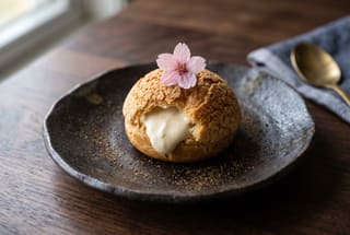 Kaze no Haru Silken Miso Cream Puff A delicate vegan cream puff with white miso filling on a dark ceramic plate, garnished with a single pink sakura blossom and tea powder.
