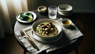 Elegant vegan mushroom miso risotto in a white bowl, with steamed greens on a dark table, simple cutlery, and soft natural lighting.