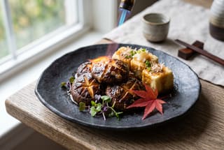 Flambéed shiitake mushrooms and agedashi tofu on a handmade ceramic plate with a flickering blue flame and shiso garnish.