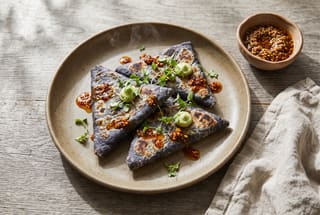 Crispy Blue Corn Tetelas with Charred Wild Mushrooms and Citrus Macha Crispy triangular blue corn tetelas topped with glossy red salsa macha, fresh herbs, and whipped avocado on a rustic ceramic plate.