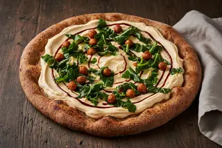 Artisan teff pizza topped with whipped peri-peri cream, Sukuma Wiki, and harissa chickpeas, drizzled with tamarind on a rustic wood table.
