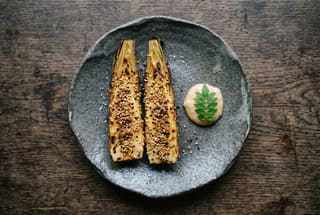Two golden-brown encrusted bamboo shoots on a dark ceramic plate with a dollop of miso cream and a green leaf on a wooden table.