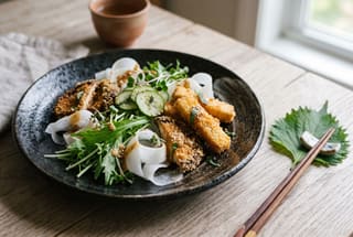 A minimalist Japanese vegan salad with breaded shiitake and tofu on a dark ceramic plate with fresh greens and daikon ribbons.