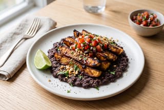 Tamarind-Glazed Cassava Wedges with Velvety Njahi Ragu Charred tamarind cassava wedges served over a creamy black bean ragu with fresh salsa and crushed peanuts on a white plate.