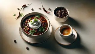 Image of a bowl of vegan espresso-infused chili topped with cilantro and vegan sour cream, beside a cup of espresso on a clean table, under warm light.