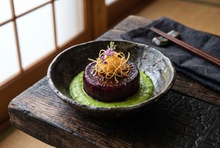 A braised daikon round in red wine sauce served on a bright green puree in a dark wabi-sabi bowl, garnished with crispy burdock.