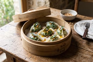 Tuscan-Style Cavolo Nero and Cashew Ricotta Dim Sum Steamed kale and cashew dumplings garnished with saffron oil and pine nuts in a bamboo steamer on a wooden table.
